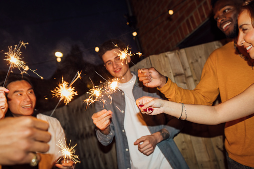 A small group of friends are at a New Year's Eve house party, they are nicely dressed and standing in a circle in the garden holding sparklers on a dark night, the garden is lit with string lights. They are celebrating the New Year in style.