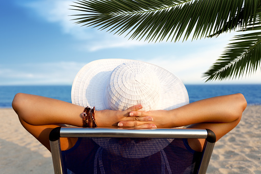 Young woman with white hat sitting on deck chair with hands beind head and relaxing at the beach, rear view. She is sunbathing by the sea and palm tree.