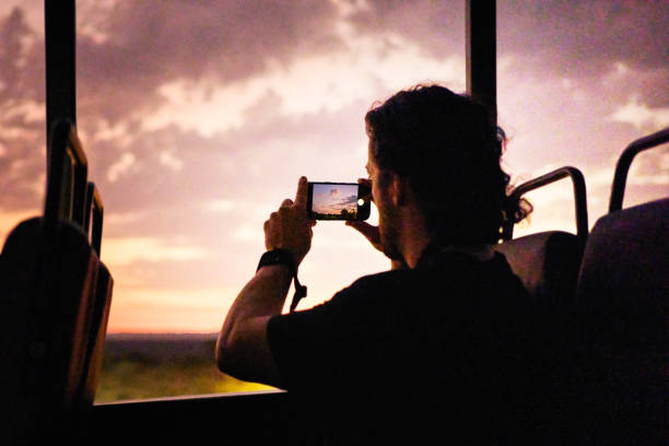 Young man taking smart phone photos of the scenery from the back of a safari vehicle driving in a wildlife reserve