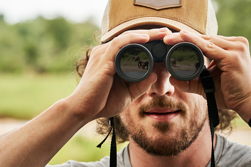 Close-up of a herd of elephants reflected in the glass of binoculars being held by a young man during a safari