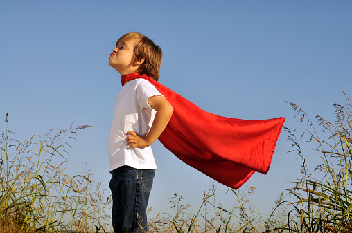 Horizontal image of a young boy standing outdoors on a clear summer day in a field of tall grass with a blue sky. He is wearing a red cape and pretending he is a superhero. The wind is blowing his cape as he considers his next mission.