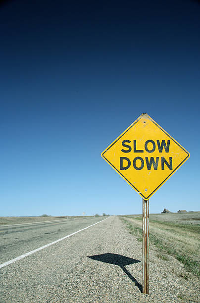 sign on flat prairie road warns drivers to slow down A yellow warning sign tells drivers to slow down. The sign casts a shadow on a paved, flat prairie road. It is a typical Saskatchewan road, beneath a beautiful blue sky, without a cloud in view. Farms can be seen in the distance. This picture is also a good reminder to all of us to slow down and enjoy life! slow stock pictures, royalty-free photos & images