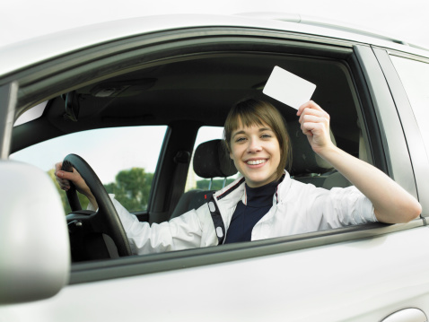 Young beautiful woman sitting in a new car and holding a blank empty sign/ driving licence/ credit card.