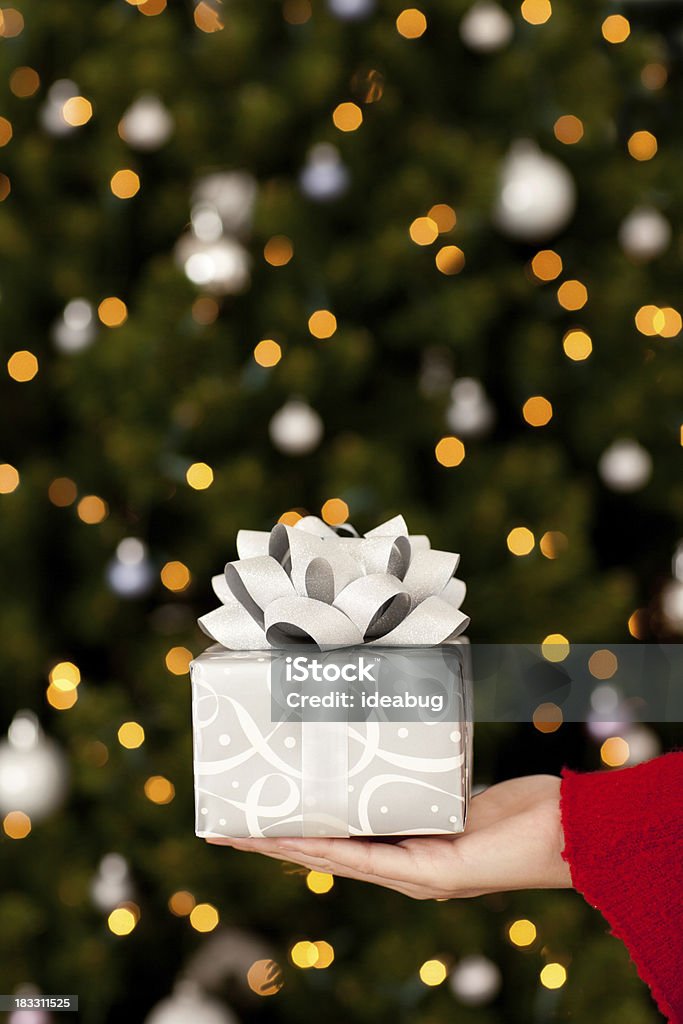 Young Woman Holding Christmas Gift with Lights and Tree Color photo of a young woman holding a small Christmas gift with Christmas tree and lights blurred behind her. Silver Colored Stock Photo