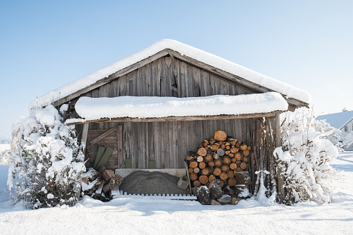 old barn in the snow during the winter day, firewood under a canopy