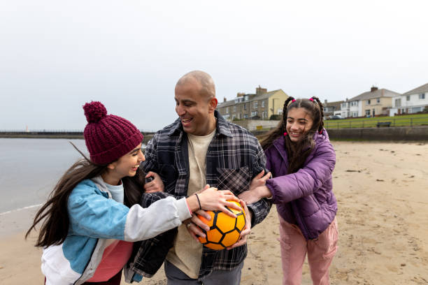 A waist-up shot of a father and his two daughters running together on a beach. They are all wearing warm clothing on a cold morning. The beach is located in Amble, Northumberland.