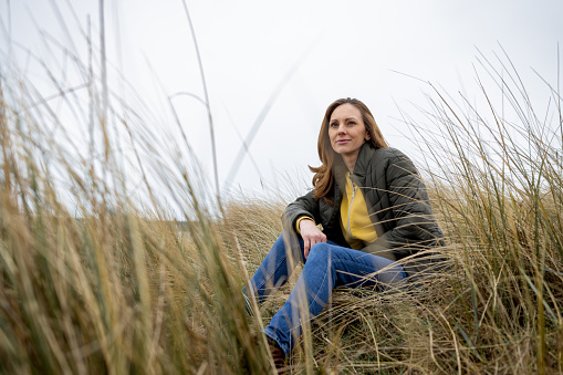 A full-length shot of a mature female adult sitting alone surrounded by marram grass. She is contemplating wearing warm clothing on a cold morning. She is located in Amble, Northumberland.
