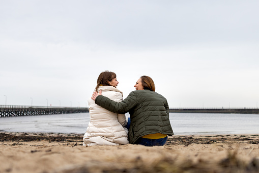 A rear view full shot of a mother and daughter sitting on a beach together. The daughter has her arm around her mother looking face to face. They are both laughing and smiling wearing warm clothing on a cold morning. They are located in Amble, Northumberland.