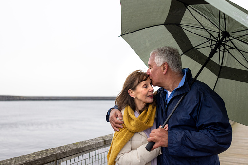 Waist-up shot of a senior couple on a pier. The male adult is kissing his wife on the head. They are all wearing warm clothing on a cold morning. The beach/pier is located in Amble, Northumberland.