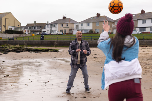 An over-the-shoulder shot of a father playing catch with her daughter on a beach along the water's edge. They are both wearing warm clothing on a cold morning. The beach is located in Amble, Northumberland.