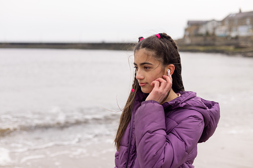 A waist-up shot of a girl walking along a seashore listening to music on wireless Bluetooth headphones. She is wearing warm clothing on a cold morning. The beach is located in Amble, Northumberland.