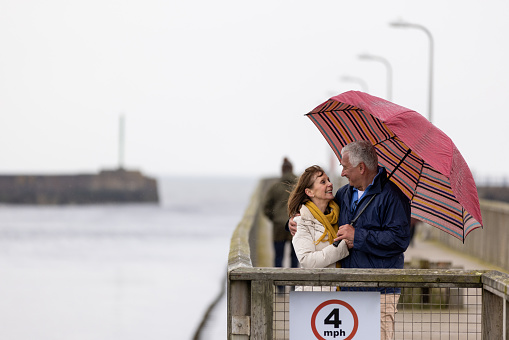 Full shot of a senior couple on a pier looking at each other lovingly under an umbrella. They are both wearing warm clothing on a cold morning. The beach/pier is located in Amble, Northumberland.