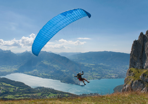 A paraglider with a bright blue sport parachute just launched from a ridge and cliff high above Annecy Lake in the French Alps.