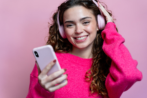 Young woman listening to music with headphones and smartphone on a pink background