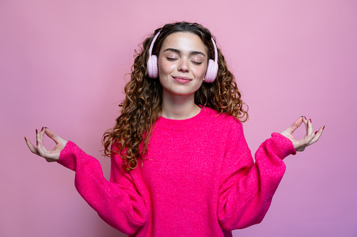 Beautiful young woman with long curly hair in pink sweater listening to music in headphones on pink background and meditating