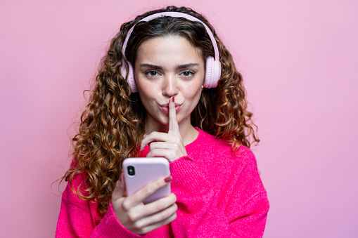 Portrait of a beautiful young woman with curly hair in pink sweater and headphones showing silence gesture on pink background.