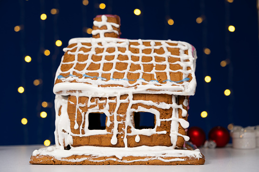 Christmas house made of gingerbread cookies decorated with sugar icing on blue background with bokeh garland.