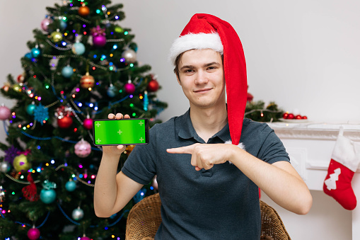Close-up portrait of a young man in a santa hat on the background of a Christmas tree, pointing at a phone with a green screen