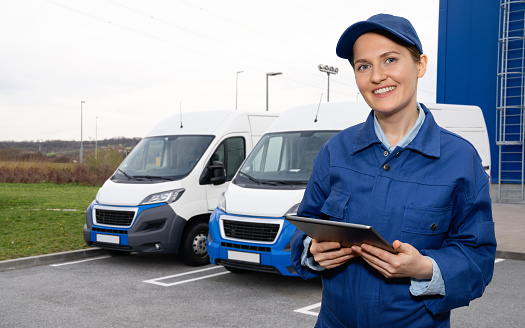 Woman worker wearing blue uniform is holding digital tablet and looking at the camera. Vans are parked in row. Vans are parked in row. Commercial fleet.