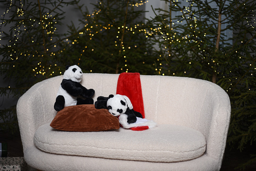 Christmas interior with green Christmas trees decorated with lights behind a white sofa where two toy pandas are sitting with a red elf hat.