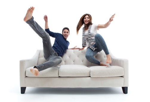 A smiling playful interracial couple jumps over the back of a living room couch.  Horizontal, isolated on white. (Slight motion blur; focus is sharp on womans face).