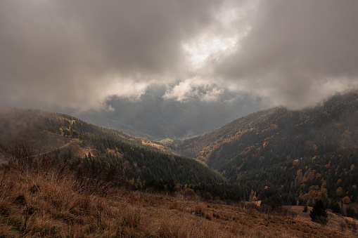 Panoramic view from above of a natural mountain environment being covered by stormy clouds and being illuminated by the sunlight, in late afternoon, in autumn. district of Paularo, F.V.G. region, Italy