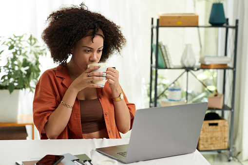 Woman with fluffy curly hair drinking cup of coffee and watching product presentation or educational video on laptop