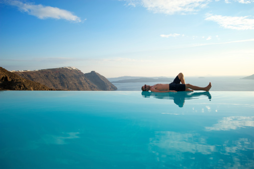 Young man reclining lying back on the edge of an infinity pool with a sweeping sea view over Santorini, Greece