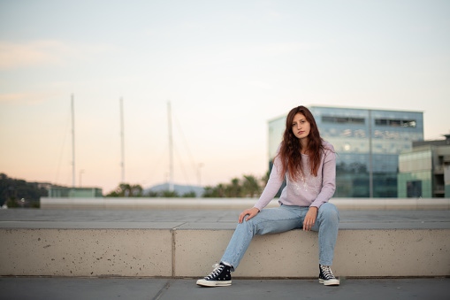 Young woman in casual clothes posing in front of an urban setting at sunrise.