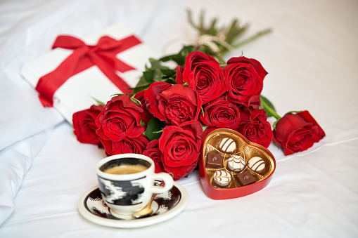 From above of composition of bunch of red roses and cup of aromatic fresh coffee with heart shaped box of chocolates placed on white soft bed in hotel room