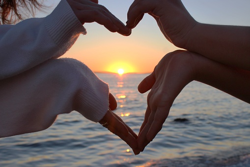hands join in the shape of a heart against the backdrop of a beautiful sunset on the sea,heart symbol  sign of the heart, connection, people’s love, trust and tenderness