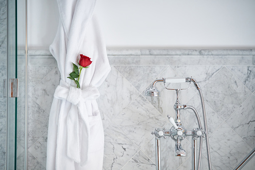 Interior of luxury bathroom with vintage faucet and white terry bathrobe with red rose placed in belt hanging on marble tiled walls