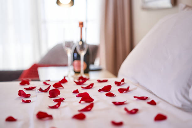 Bed with white bedclothes covered with rose petals From above closeup of bed covered with white bedclothes and cushions with scattered petals of fresh red roses and bottle of champagne with glasses on bedside table in hotel room bed decoration stock pictures, royalty-free photos & images