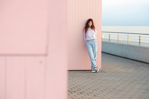 Young woman posing in front of the sea leaning against a pink wall.