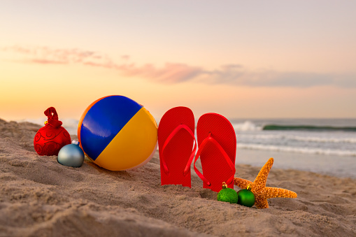 This is a conceptual photograph symbolizing taking a Christmas Holiday vacation to the beach. There are colorful flip flops stuck upright in the sand. There is a beautiful sunrise in the background.