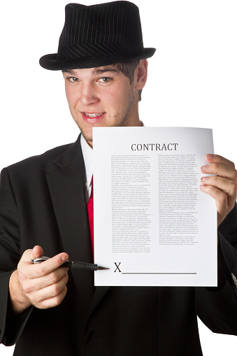 Smiling man with a cheesy grin gesturing for you to sign a contract, isolated on white. Man is mid 20s caucasian, is clean shaven with brown hair and is wearing a blue suit coat, shirt and red tie and fedora. He is portraying a role as a used car salesman or some other type of high pressure sales.