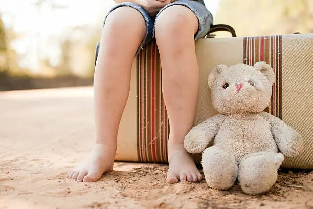 Color photo of a 6-7 year old girl sitting barefoot alone on a vintage suitcase with an old, raggedy teddy bear on a dirt road.
