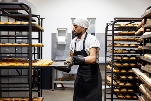 Latin American baker taking trays of bread out of the oven at an industrial bakery - food processing plant concepts
