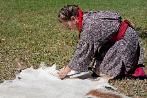 A French and Indian War reenactor portraying an Algonquin Indian woman in period costume.  She is scraping deer hide that will be later used as shelter protection.For similar photos check out my