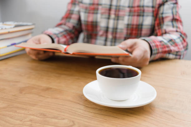 istock Cup of coffee on wooden table, unrecognizable man reading book, studying or working, sitting at the desk with stack of books, focus on foreground 1833506731