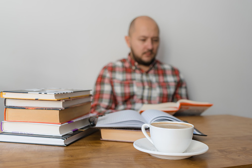 Man reading a book, studying or working, sitting at the table with stack of books and cup of coffee, focus on foreground.