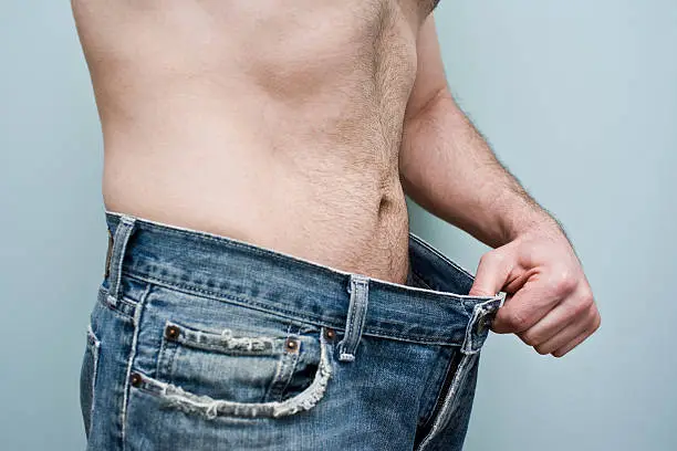 Man stands with a pair of oversize blue jeans focussing on his weight loss