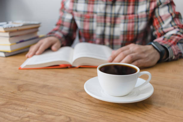 istock Cup of coffee on wooden table, unrecognizable man reading book, studying or working, sitting at the desk with stack of books, focus on foreground 1833508861