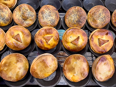 Horizontal high angle closeup photo of a variety of freshly baked traditional quality Australian pies: cheese and bacon, beef and red wine, lamb and rosemary and chicken and vegetable, hot out of the oven in metal baking tins oin a Cafe Bakery. Armidale, New England high country, NSW.