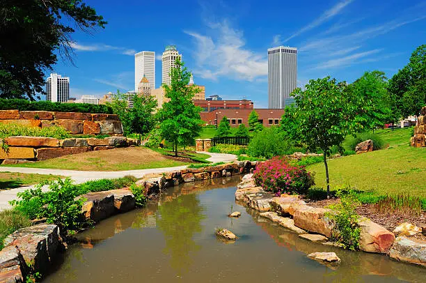 "Tulsa downtown skyline from a park with trees, grass, rocks, and a stream in the foreground."