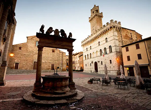 "An ornate well silhouette stands in the foreground of the town plaza in Montepulciano, Italy. In the background, the prominant Town Hall tower is starting to be illuminated with pre-dawn light."