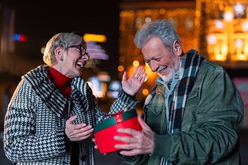 Caring cheerful senior woman giving Christmas present to a smiling senior man on the street. Elderly man surprised with a gift from his old friend. Holiday season concept.