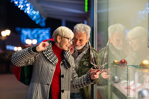 An elderly couple is shopping for Christmas gifts for friends and family, standing in front of a store window and discussing things to buy.
