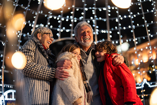 Overjoyed senior couple having fun with granddaughter and grandson surrounded with Christmas lights during winter holidays.