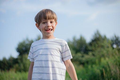 Cute happy boy in light T-shirt without front tooth smiles on summer day in nature. Change of teeth.
